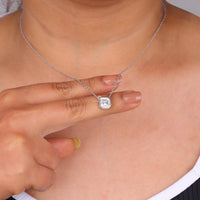 Person wearing a silver necklace with a pendant, pointing to it, Elegant Bezel Set Moissanite Jewelry, Sustainable Moissanite Anniversary Necklace, Conflict Free Asscher Cut Wedding Jewelry