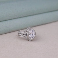 Silver ring with a large central gemstone and smaller stones on a light gray surface, Oval Moissanite Engagement Ring & Enhancer Guard – Halo Split Shank Wedding Set