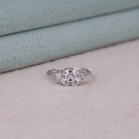 Diamond ring on a light gray surface with a neutral background, Asscher Cut Moissanite Engagement Ring – Triangle Side Stone Three Stone Design