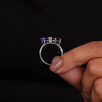 Silver ring with purple and green gemstones held by a hand against a black background