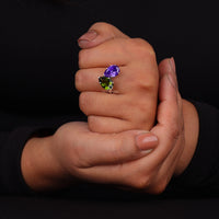 Hand wearing a ring with purple and green gemstones against a black background