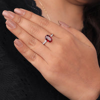 Hand wearing a ring with a red gemstone against a blurred background, Deep Red Cubic Zirconia Engagement Jewelry, Affordable Garnet Oval Promise Ring, Elegant Red Gemstone Bridal Jewelry