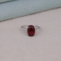 Ring with a red gemstone and diamond-studded band on a light gray surface, Classic Oval Garnet Style Engagement Ring, Glamorous Red Gemstone Hidden Halo Jewelry