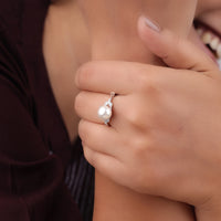 Close-up of a hand wearing a pearl ring with a blurred background, delicate pearl ring, pearl engagement ring alternative, white pearl and Moissanite ring, gold leaf ring with Moissanite, Moissanite wedding ring with pearl, eco-friendly Moissanite ring, Moissanite ring for her, feminine Moissanite ring, pearl minimalist band, two-stone pearl Moissanite ring, floral Moissanite ring, promise ring with pearl and Moissanite
