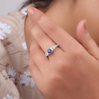 Close-up of a hand wearing a silver ring with a blue gemstone, blurred floral background, Sapphire & White CZ Art Deco Ring – Cushion and Asscher Cut Three Stone Engagement Ring