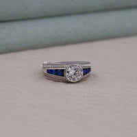 Silver ring with a large central gemstone and smaller blue stones on a neutral background