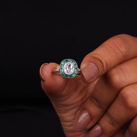 Silver ring with green and red gemstones held between fingers against a black background