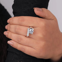 Hand wearing a silver ring with a large diamond and smaller stones on a neutral background, Sustainable Moissanite Engagement Jewelry, Ethical Moissanite Wedding Ring, Dainty Bypass Moissanite Ring