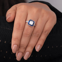 Close-up of a hand wearing a diamond ring with a blue gemstone on a black background, asscher cut sapphire wedding ring, art deco target engagement ring, milgrain bezel set sapphire ring,