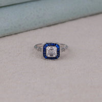Silver ring with a blue gemstone and diamond on a light gray surface, asscher sapphire anniversary ring, milgrain bezel vintage wedding band, sapphire target style engagement ring