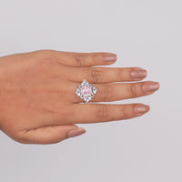 Hand wearing a ring with a pink gemstone on a light gray background, pink cushion stone promise ring for her, halo design cocktail ring for parties, cz halo statement jewelry for women