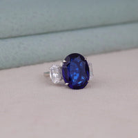 Ring with a blue gemstone and diamond on a light gray surface, three stone promise ring, oval cut sapphire engagement ring, blue gemstone anniversary ring, affordable engagement jewelry
