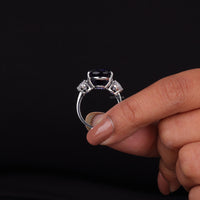 Silver ring with a dark gemstone held by a hand against a black background, Oval CZ and Blue Sapphire Trio Stone Engagement Ring, Bridal Jewelry