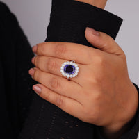 Hand wearing a ring with a purple gemstone on a gray background, Cushion Cut Blue Sapphire Engagement Ring | Halo Style CZ Wedding Jewelry