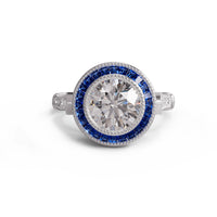 Diamond ring with blue gemstones on a white background, A ring featuring a central cubic zirconia (CZ) stone surrounded by a halo of smaller CZ stones set in a target-shaped pattern, with a metallic band and sparkling stones.