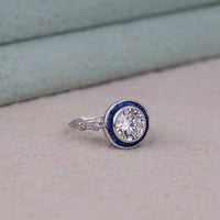 Silver ring with blue and white gemstone on a light gray surface, Round Blue Sapphire Halo Ring – Art Deco Vintage Target CZ Statement Ring