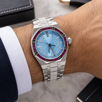 A silver stainless steel wristwatch with a textured light blue dial and a bezel accented by vivid magenta-purple stones, shown worn on a person's wrist with a white dress shirt cuff visible.