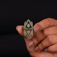 Hand holding a decorative ring with green and white gemstones against a black background