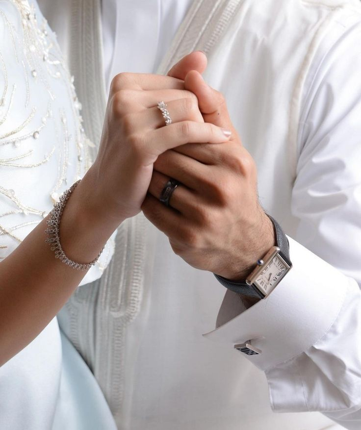 Close-up of hands holding each other with wedding rings, wearing formal attire.