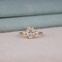 Oval cut Moissanite engagement ring with bezel-set 10.00 × 7.00 mm center stone, pear cut side stones, elegant yellow gold finish, and minimalist three-stone design.