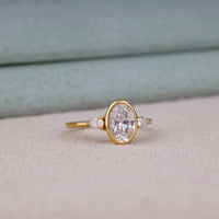 Oval cut Moissanite engagement ring with bezel-set 10.00 × 7.00 mm center stone, pear cut side stones, elegant yellow gold finish, and minimalist three-stone design.