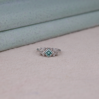 Princess cut moissanite engagement ring with aqua green center stone, leaf accent side stones, and nature-inspired design